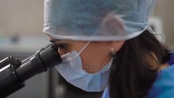 Inyección de cerca de una doctora en una cara que examina bajo un microscopio gotas de sangre para detectar enfermedades y confirmar el estado saludable de los pacientes — Vídeo de stock