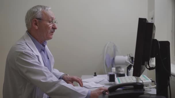 Взрослый врач в халате делает анамнез пациента частной клиники, чтобы сделать результаты в медицинской карте единой компьютерной базы поликлиники — стоковое видео