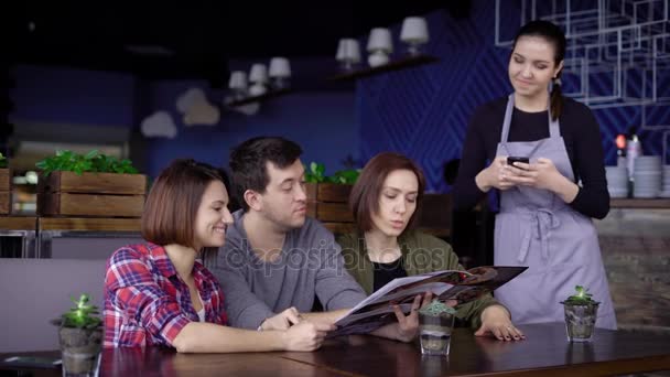 La compañía de amigos adultos hace un pedido para el menú en el restaurante, refiriéndose a la camarera, los amigos eligen la cena, sentado en una mesa de madera — Vídeos de Stock