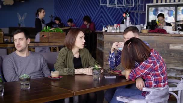 Τέσσερις φίλοι κάθονται σε ένα ξύλινο τραπέζι σε ένα καφέ και καφέ, συζητούν επιχειρηματικά σχέδια σε μια χαλαρή ατμόσφαιρα, με ένα φλυτζάνι καπουτσίνο — Αρχείο Βίντεο