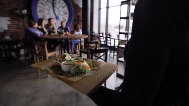 Bir kadın garson bir ahşap masanın üzerine, arkasında oturan bir grup erkek ve kadın, sebze aperatifler ve ekşi krema sosu ile bir tepsi koyar
