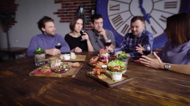 En la mesa de madera en el restaurante hay un montón de bocadillos vegetarianos, en el fondo una compañía de amigos está sentada que bebe vino tinto y se lo pasa bien juntos — Vídeos de Stock