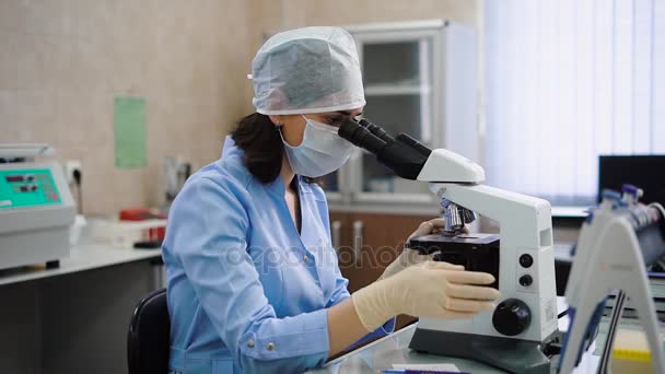 Trabajadora de laboratorio usando microscopio. Especialista femenina joven en uniforme sentada a la mesa y utilizando microscopio . — Vídeo de stock