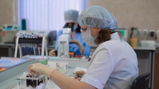 Vrouwelijke specialist controle bloed monster. Jonge vrouw in uniforme zittend aan tafel en kijken door exemplaar met bloed aan klinisch laboratorium. — Stockvideo