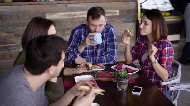 Compagnie d'amis assis à la table dans le café profiter de leur repas et de parler entre eux. Deux hommes et deux femmes discutent en déjeunant au restaurant. Les gens boivent et mangent ensemble . — Video