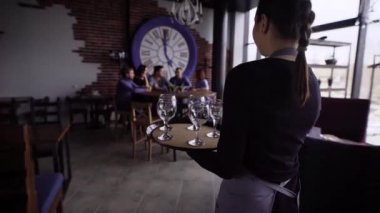 Tabloya yürüyüş ve misafirler için bir tepsi ile gözlük şarap için taşıyan genç misafirperver garson. Parti restoranda oturan ve onların içecekler için bekleyen insanlar.