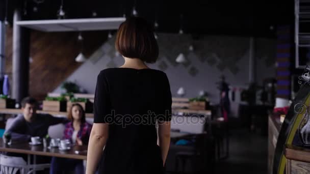 Wizerunek Szczupła młoda kobieta w czarnej sukni, spacer do kawiarni do jej przyjaciół. Firma osób siedzących stołu powitaniem przybyłych kobiece kobieta kolega obejmując i całując ją serdecznie. — Wideo stockowe
