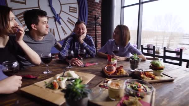 Cinco amigos están sosteniendo copas de vino tinto y sentados a la mesa servidos con diferentes aperitivos en el restaurante y haciendo fiesta. Hombres y mujeres están pasando tiempo libre en la cafetería cerca de la ventana . — Vídeo de stock