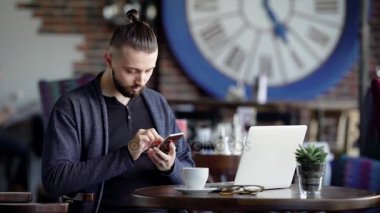 Genç çekici hippi bir kafe, modern iç. Bir adam kahve ile diz üstü yanındaki bir smartphone kullanarak bir masada oturuyor.
