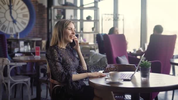 Mujer joven y feliz hablando por teléfono y usando la computadora en una cafetería. Una hermosa chica, se comunica emocionalmente por teléfono con su amigo . — Vídeos de Stock