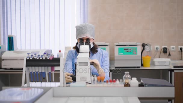 Asistente de investigación femenina en uniformes azules, máscara protectora, gorra y guantes sentada en laboratorio examinando muestras médicas usando microscopio. Mujer joven en uniforme trabajando con el espécimen y tomar notas . — Vídeos de Stock