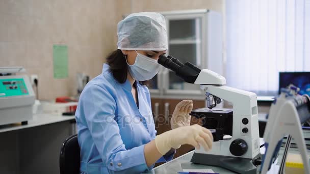 Assistente de laboratório profissional feminino em jaleco azul, máscara protetora, boné e luvas sentadas no armário e examinando amostras médicas sob o microscópio . — Vídeo de Stock