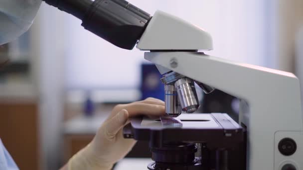 Primo piano del microscopio da laboratorio per l'ingrandimento e la ricerca di campioni medici. Immagine di una mano di assistente che esamina un piccolo bicchiere con un campione attraverso la lente di attrezzature speciali . — Video Stock