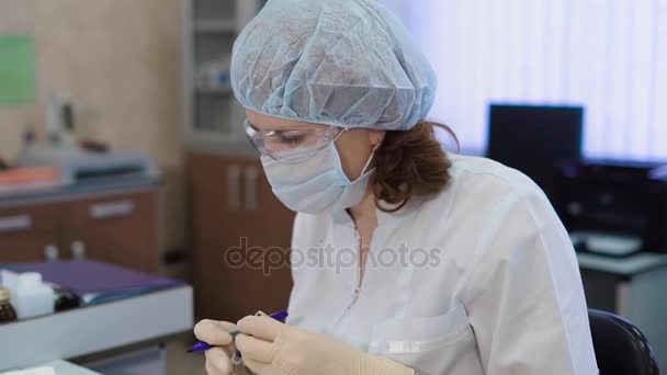 Forschungsassistentin in weißem Laborkittel, Schutzmaske, Mütze und Handschuhen sitzt im medizinischen Labor und arbeitet mit Reagenzgläsern mit Blutproben. Krankenschwester sortiert Proben und macht Notizen. — Stockvideo