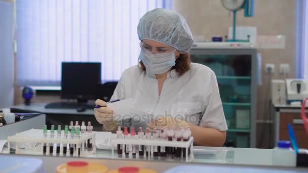 Forschungsassistentin in weißen Laborpeelings, Schutzbrille, Maske, Mütze und Handschuhen sitzt am Tisch im medizinischen Labor mit dem Satz Reagenzgläser und bereitet Blutproben für die Untersuchung vor. — Stockvideo