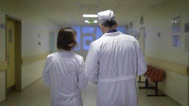 Erkek ve kadın hekimlerin arkadan görünüm koğuş koridordan yürürken ve Birbirinizle konuşmak laboratuvar mont giymiş. Üniformalı tıp profesörü ile onun meslektaşınla laboratuvara gidiyor.