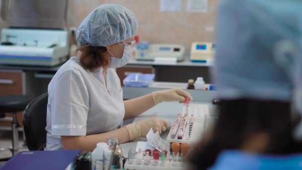 Assistente di ricerca vestita con camice bianco da laboratorio, occhiali protettivi, maschera, berretto e guanti seduti a tavola con una rastrelliera di provette che preparano campioni di sangue da esaminare in laboratorio medico . — Video Stock