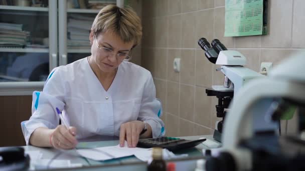 Assistente de pesquisa feminina profissional em casaco branco sentado à mesa com microscópio e calculadora na frente de seu processamento de dados médicos. Doutor em uniforme que trabalha com resultados do exame . — Vídeo de Stock