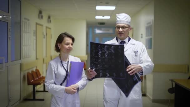 男性と女性の医師は病棟の廊下を歩くと roentgenograms を議論するに実験用の白衣に身を包んだ。制服の医学部教授は、x 線撮影を保持しています。 — ストック動画