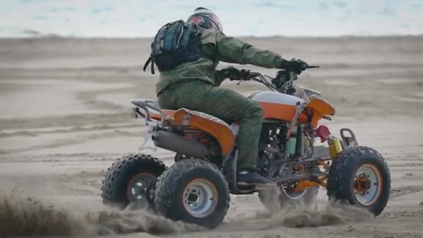Человек в шлеме на голове выполняет экстремальный трюк на спортивном квадроцикле на пляже, песок вылетает из-под колес автомобиля — стоковое видео