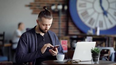 Yetişkin bir adam modern bir kafede kahve içer, kuyruğunu sıkışmış bir saçlı bir adam internette son haberler cep telefonundan taradığında, masanın üzerinde bir dizüstü bilgisayar var