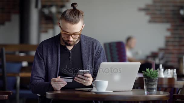 Młody człowiek siedzi w kawiarni i sprawia, że zamówienie online stylowe ubrania w sklepie internetowym, osoba korzysta z karty kredytowe i telefon komórkowy dla płatności, na biurku jest komputer przenośny — Wideo stockowe