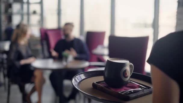 De ober draagt een grote cappuccino-kop voor het jonge paar in de lade, die een aperitief in een restaurant uitvoert, een man en een vrouw praten over het afgelopen weekend — Stockvideo