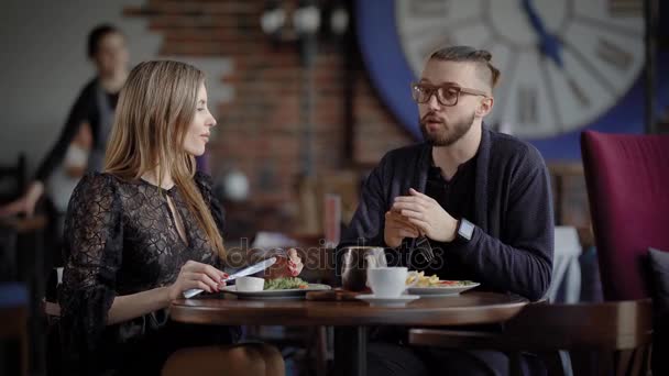 Um jovem casal de amantes comeu uma refeição saudável em um grande café, um homem com má visão e um relógio inteligente em suas mãos conta uma história divertida para uma mulher bonita. — Vídeo de Stock