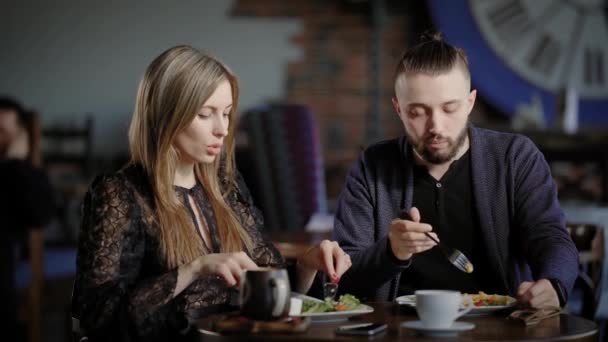 Una pareja joven comiendo una ensalada en un restaurante, un hombre y una mujer bonita hablando durante una comida, sobre la mesa hay tazas de café — Vídeos de Stock