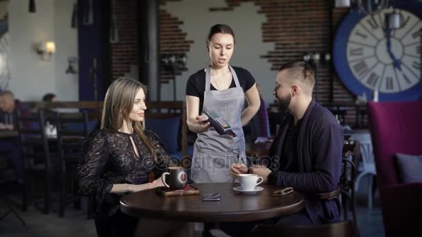 Een jong koppel betaalt een factuur voor het diner aan een ober in een restaurant met een bankkaart, een man en een vrouw drank cappuccino en americano in kopjes — Stockvideo