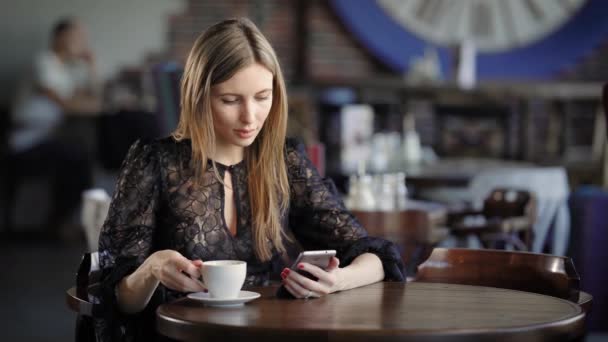 Donna elegantemente vestita beve caffè in un piccolo ristorante, seduto a un tavolo di legno su cui c'è una tazza, la bellezza corrisponde a suo marito su un telefono cellulare. — Video Stock