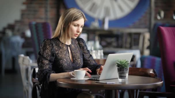 Femme d'affaires communique avec ses collègues au travail, assis dans un petit restaurant, une femme boit du café et imprime du texte sur un ordinateur portable portable qui l'aide à travailler à distance — Video