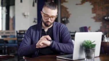 Restoranda oturan, seyir ve smartwatch dokunmadan sakallı hippi yakın çekim. Erkek Tasarımcısı eline istemcisi dizüstü bilgisayar ve modern kol saati kafeteryada masasında bekliyor.