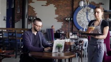 Misafirperver genç garson kafede kahve cam istemciye getiriyor. Rahat kıyafetler şık genç adam restoranda boş vakit ve laptop ile çalışma oturuyor.