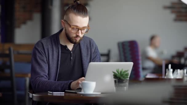 Primer plano de hombre guapo en anteojos, camiseta negra y chaqueta azul sentado en la cafetería con un portátil moderno. Escritor masculino trabajando como freelancer en oficina móvil escribiendo texto en gadget digital . — Vídeo de stock