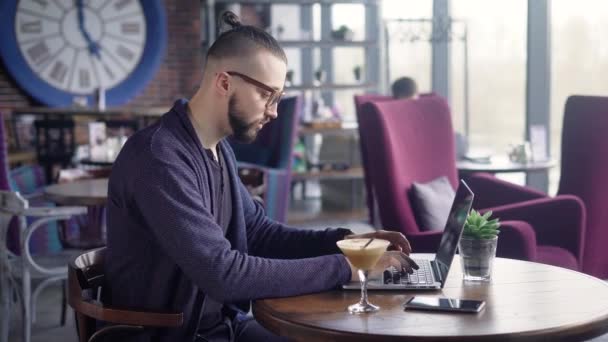 Side view of a man in black t-shirt and blue jacket sitting at the table using laptop. Young hipster with modern eyeglasses typing text on PC. Popular blogger writing article in social networks. — Stock Video