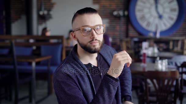 Portret van bebaarde hipster in stijlvolle bril zittend in restaurant en kijken naar de camera. Knappe man aan de tafel in het cafetaria met mensen op de achtergrond is zijn bril opstijgen. — Stockvideo