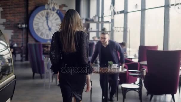 Acercamiento de una delgada mujer de pelo largo caminando a través de la cafetería hasta la mesa con su amigo varón. El hombre está de pie saludando a la dama que viene. Pareja sentada en restaurante hablando y sonriendo la una a la otra . — Vídeos de Stock