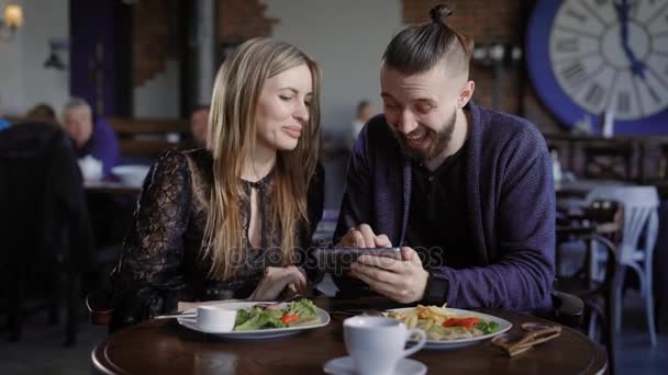 Счастливая пара обедает вместе и смотрит на смартфон. Мужчина и женщина с помощью цифрового гаджета смеются над изображениями внутри. Друзья в ресторане смотрят на дисплей мобильного телефона и улыбаются . — стоковое видео