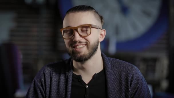 Retrato de hipster positivo bonito olhando para a câmera e sorrindo. Homem elegante de casaco azul e óculos elegantes olhando diante dele tentando ser sério, removendo os olhos e rindo . — Vídeo de Stock