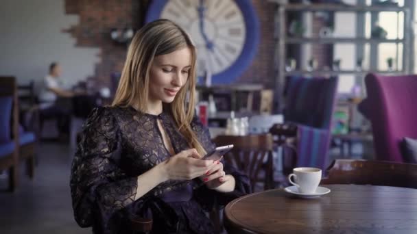 Primer plano de la mujer de negocios vestida con vestido negro está sentado en la cafetería con un teléfono inteligente moderno. Adorable dama se relaja en la mesa durante el almuerzo escribiendo texto en el dispositivo digital y sonriendo . — Vídeos de Stock