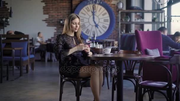 Femme d'affaires en robe noire profitant de la pause déjeuner assis dans un café avec des gens en arrière-plan et en utilisant un smartphone moderne. Adorable dame se détendre au restaurant en regardant l'appareil numérique et souriant . — Video