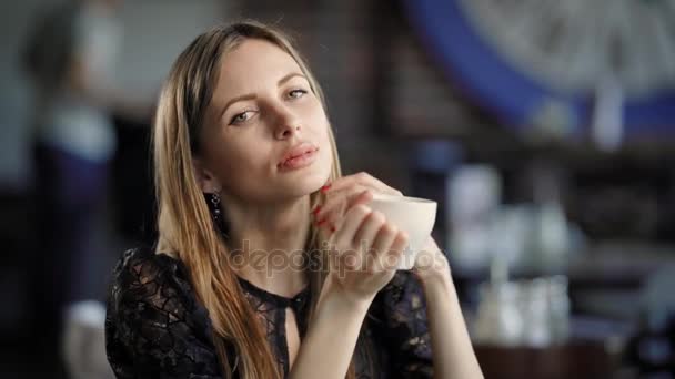 Vacker kvinna sitter vid bordet i restaurangen med suddig bakgrund och hålla kopp kaffe. Bedårande dam med naturlig make-up i svart spetsklänning tittar på kameran och leende. — Stockvideo