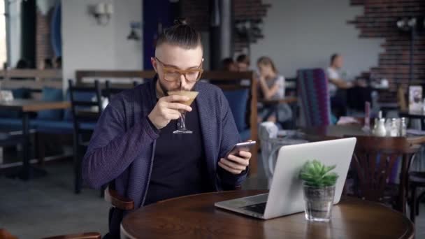 Мужчина пользуется телефоном, сидя в кафе. Молодой человек использует телефон, сидя в кафе с напитками и ноутбуком . — стоковое видео