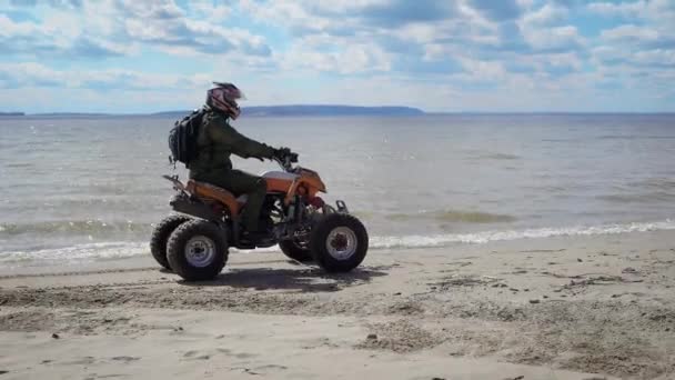 Hombre vestido con equipo de protección y casco a caballo ATV y conducir a lo largo de la orilla del mar con alta velocidad. Aficionado al turismo extremo con mochila detrás corriendo en su quad bike en la playa . — Vídeo de stock