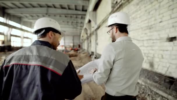 De klant in een beschermende helm controleert het werk van bouwers op het object in aanbouw, mensen lopen door het gebouw van de oude fabriek — Stockvideo