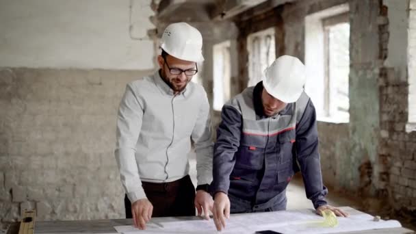 Взрослый инженер объясняет рабочему новый строительный план, который был спроектирован архитектором, люди находятся в заброшенном здании — стоковое видео
