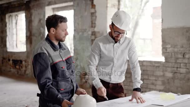 En affärsman i en hjälm på huvudet och en professionell konstruktion diskuterar ritningar av byggnaden, männen är på byggarbetsplatsen — Stockvideo
