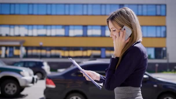 Зайнята бізнес-леді говорить на своєму мобільному телефоні, тримаючи важливі контракти, вона йде на парковці, де стоять автомобілі — стокове відео