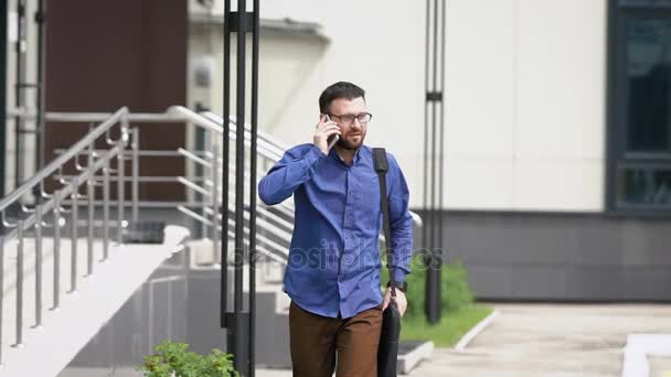 Slow motion van een drukke man die onderaan de straat loopt en spreekt op zijn mobiele telefoon met een zakenpartner, op zijn schouder een zak van documenten — Stockvideo